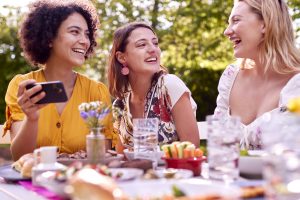 Three Female Friends Looking At Photos On Mobile Phone Eating Meal Outdoors In Summer Garden At Home